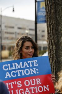 woman holds red-&-blue sign with white text reading “Climate Action: It’s Our Obligation”.