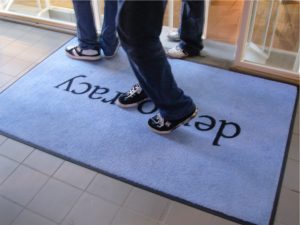 The focus is on doormat on a tile floor; a doorway is visible above it. The doormat has the word “democracy” inscribed; 2 pairs of lower legs in jeans and sneakers stand on the mat; another is standing in the doorway.