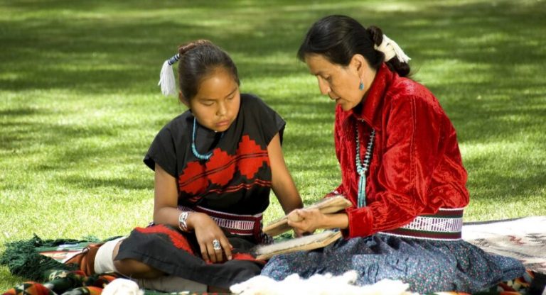 Native American women, with their hair tied back with feather and fabric, wearing black and red clothing, with beaded belts and silver and turquoise jewellry read together while sitting on blanket on sun-dappled lawn